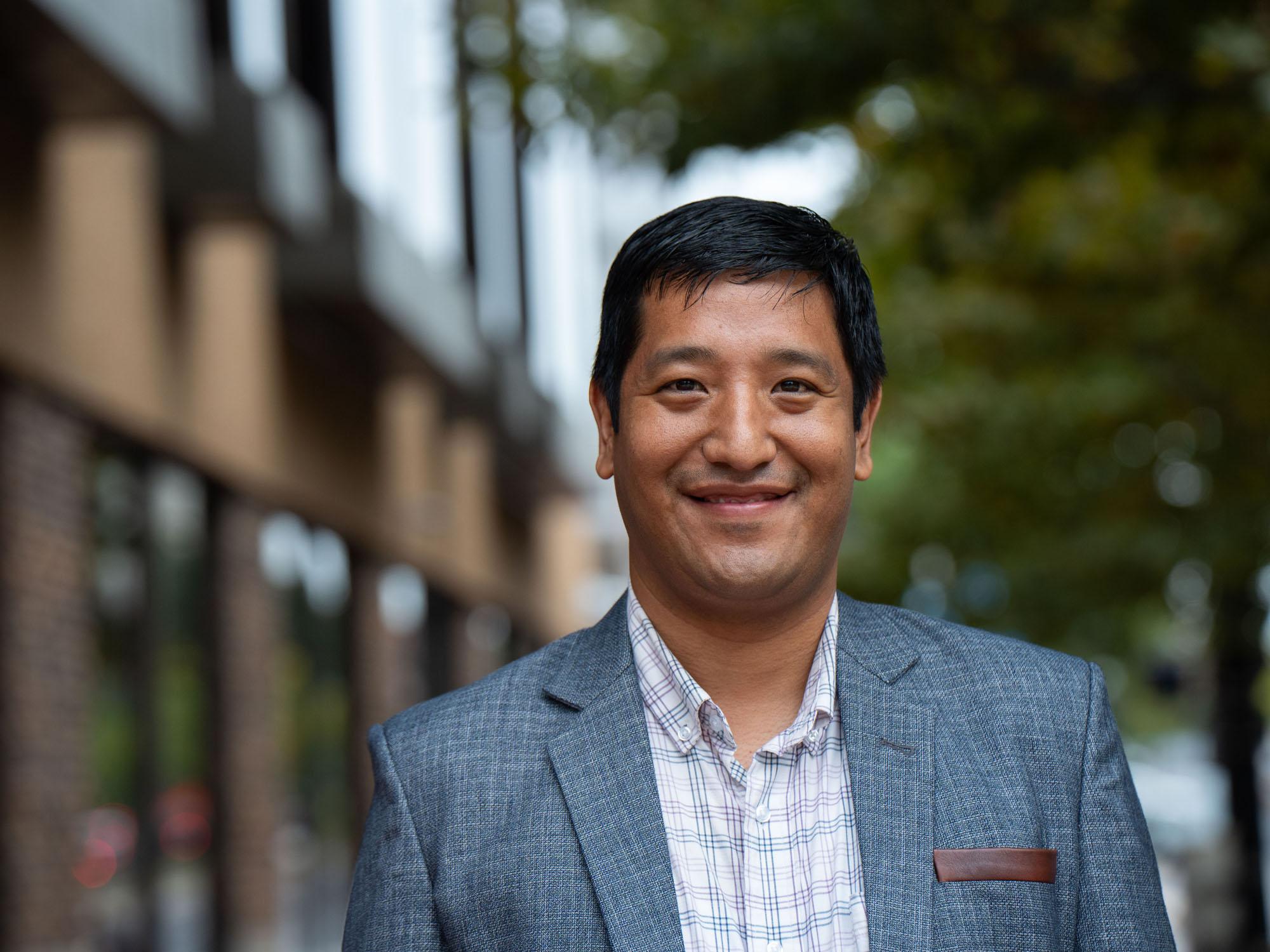 Nivel Gurung poses, smiling, at the camera, while standing on the UBalt campus outside its Academic Center.